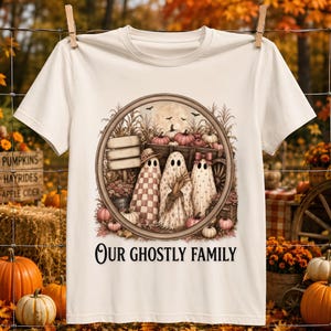Peut inclure: Un t-shirt couleur crème avec une illustration circulaire représentant trois fantômes dans un champ de citrouilles, un chariot en bois, des tiges de maïs et une pleine lune. Le texte "OUR GHOSTLY FAMILY" est imprimé en noir sous l'image.
