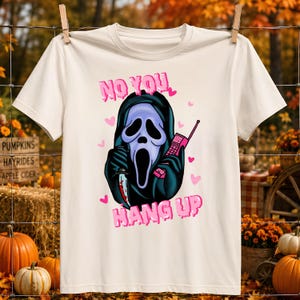 Può includere: Una t-shirt a maniche corte color crema con la grafica di una figura mascherata in una tunica nera con cappuccio, che tiene in mano un coltello e un telefono vintage rosa. Il testo "NO YOU HANG UP" è stampato in rosa con piccoli cuori.