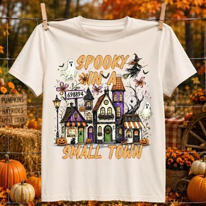 May include: A cream-colored short-sleeve t-shirt featuring a graphic print of a spooky town with houses, pumpkins, ghosts, and a witch. The text reads "SPOOKY IN A SMALL TOWN" and includes a sign with the number "698894" in the center of the design.