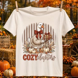 Puede incluir: Una camiseta de manga corta color crema con un gráfico de un ganso blanco que lleva un cárdigan beige, gafas y una pajarita roja. El ganso sostiene una pila de libros. Debajo, el texto dice COZY chapters, rodeado de calabazas.