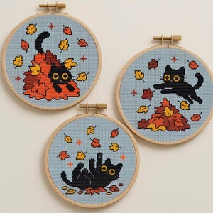 May include: Three cross-stitch hoops showing black cats amongst autumn leaves. The cats are surrounded by falling leaves in orange, yellow, and red. The background is a light blue fabric, and the hoops are made of wood.