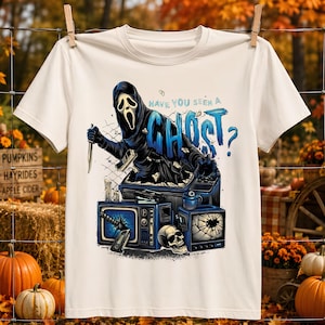 May include: A cream-colored t-shirt featuring a graphic of a hooded figure with a white mask holding a knife, emerging from a television set. The text reads "HAVE YOU SEEN A GHOST?" above vintage televisions, a skull, and a cassette tape.