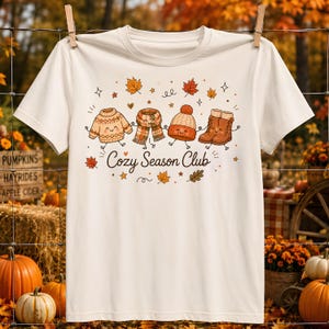 May include: A cream-colored short-sleeve t-shirt featuring a graphic of a sweater, scarf, beanie, and boots holding hands. The text "Cozy Season Club" is printed in brown script below the characters, surrounded by small autumn leaf illustrations.