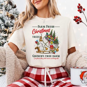 May include: A cream-colored t-shirt featuring a graphic of a green character sitting on a sleigh filled with gifts and small trees. The text reads Farm Fresh Christmas Trees, Grinch's Tree Farm, and Complimentary hot cocoa and beignets.
