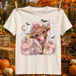 May include: A cream-colored short-sleeve t-shirt featuring a graphic of a Highland cow wearing pink sunglasses and a bow, blowing a bubblegum bubble. The cow is surrounded by several decorative pumpkins, small bats, and a crescent moon.