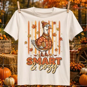 May include: A cream-colored t-shirt featuring a graphic of a goose wearing a beanie, glasses, and a cardigan, holding a book labeled Autumn Notes. The design includes falling maple leaves, vertical stripes, and the text SMART & cozy.