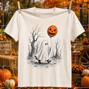 May include: A cream-colored short-sleeve t-shirt featuring a black and white ink-style illustration of a ghost walking through a forest of bare trees while holding an orange balloon with a carved jack-o'-lantern face printed on the front.