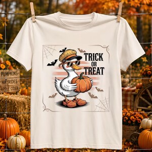 Op de afbeelding: Een crèmekleurig t-shirt met een afbeelding van een witte eend met een beige pet en zonnebril, die een oranje pompoen vasthoudt. Het ontwerp bevat de tekst "TRICK OR TREAT" in het zwart, omringd door kleine zwarte vleermuizen en spinnenwebben.