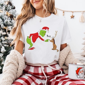 Puede incluir: Una camiseta blanca de manga corta con un gráfico de un personaje verde con traje de Papá Noel rojo tocando la nariz de un perro marrón con astas. El texto Boop está impreso en negro sobre el perro. Se lleva con pantalones de cuadros rojos.