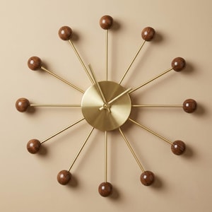 May include: A wall clock featuring a central circular brushed brass face with matching brass hands. Twelve thin brass spokes radiate outward from the center, each tipped with a polished dark brown wooden sphere, creating a sunburst design.
