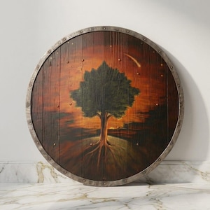 May include: A circular wooden shield featuring a painted scene of a dark green tree with visible roots against a sunset-coloured orange and brown sky. The shield has a metallic silver-toned rim secured with small rivets around the entire perimeter.