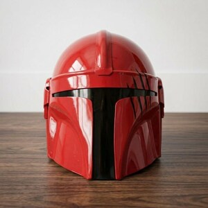 K&ouml;nnte beinhalten: Ein gl&auml;nzender roter Helm mit einer glatten, abgerundeten Kuppel und einem dunklen, horizontalen Visier. Die Vorderseite weist auf der rechten Seite eine Reihe d&uuml;nner, schwarzer, vertikaler Kratzspuren auf, die dem Finish ein abgenutztes Detail verleihen.