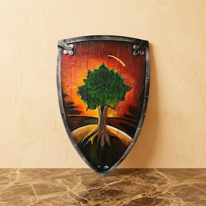 Pode incluir: Um escudo com borda metálica escura e rebites. A superfície de madeira apresenta uma cena pintada de uma árvore verde com tronco castanho, contra um céu vibrante de pôr do sol laranja e vermelho com uma lua crescente amarela acima.