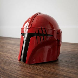 K&ouml;nnte beinhalten: Ein gl&auml;nzender roter Helm mit einer glatten, abgerundeten Kuppel und einem T-f&ouml;rmigen schwarzen Visier. Die Vorderseite des Helms weist eine Reihe von f&uuml;nf markanten, vertikalen schwarzen Kratzspuren auf der rechten Seite auf.