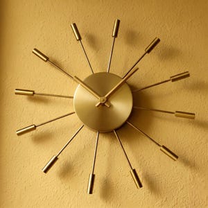 Puede incluir: Un reloj de pared dorado con un diseño de rayos de sol. El reloj presenta una esfera circular con manecillas y varillas radiales que terminan en puntas cilíndricas. Está montado en una pared texturizada de tono amarillo.