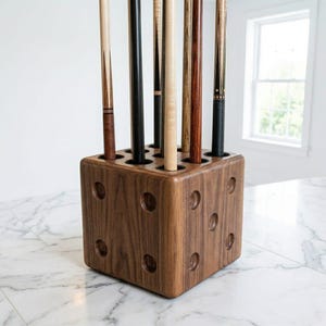 May include: A wooden pool cue holder in a cube shape, with circular cutouts on the sides. The holder is filled with several pool cues of varying colors and designs, including black, brown, and natural wood tones. The holder sits on a marble surface.