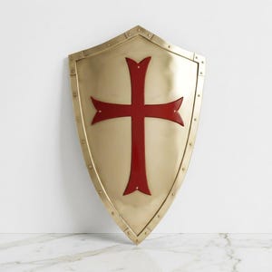 May include: A polished gold-tone metal heater shield featuring a prominent deep red cross emblem centered on its surface. The shield is constructed with a reinforced border secured by small rivets along the entire perimeter of the pointed shape.