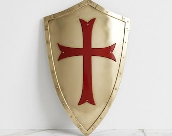 Può includere: Uno scudo in metallo color oro lucido con un prominente emblema a croce rosso scuro centrato sulla superficie. Lo scudo è realizzato con un bordo rinforzato fissato da piccoli rivetti lungo l'intero perimetro della sua forma a punta.