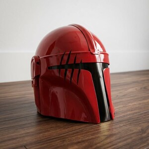 K&ouml;nnte beinhalten: Ein gl&auml;nzender roter Helm mit einem T-f&ouml;rmigen schwarzen Visier und einer Reihe von f&uuml;nf vertikalen, krallenartigen schwarzen Kratzspuren im oberen vorderen Bereich. Der Helm hat eine glatte, abgerundete Kuppel und einen markanten Mittelsteg.