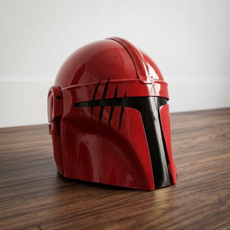 K&ouml;nnte beinhalten: Ein gl&auml;nzender roter Helm mit einem T-f&ouml;rmigen schwarzen Visier und einer Reihe von f&uuml;nf vertikalen, krallenartigen schwarzen Kratzspuren im oberen vorderen Bereich. Der Helm hat eine glatte, abgerundete Kuppel und einen markanten Mittelsteg.