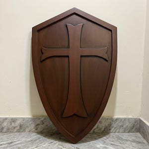 May include: A dark brown wooden shield featuring a pointed top and a tapered bottom. The shield has a raised, centered cross design with flared ends. The wood grain is visible throughout the surface, and the edges are framed by a raised border.