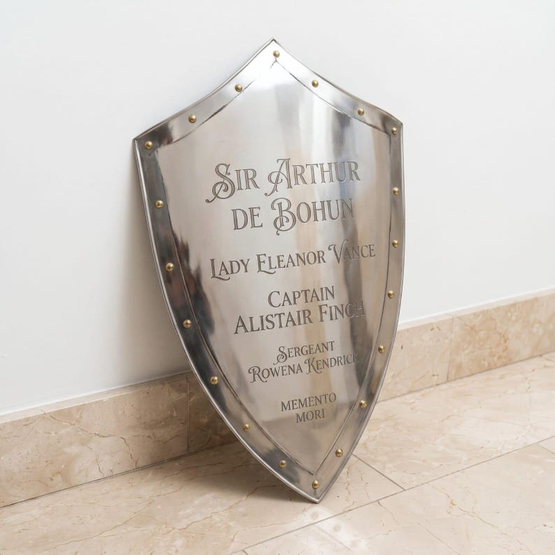 Pu&ograve; includere: Uno scudo in metallo color argento lucido con base a punta e rivetti dorati lungo il perimetro. La superficie &egrave; incisa con i nomi Sir Arthur de Bohun, Lady Eleanor Vance, Captain Alistair Finch, Sergeant Rowena Kendrick e Memento Mori.