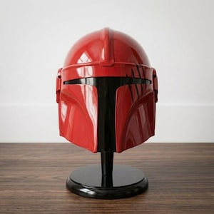 K&ouml;nnte beinhalten: Ein gl&auml;nzender roter Helm mit einem T-f&ouml;rmigen schwarzen Visier und einem Mittelsteg, der von vorne nach hinten verl&auml;uft. Der Helm weist auf der rechten Seite schwarze kratzerartige Markierungen auf und steht auf einem runden schwarzen Sockel.