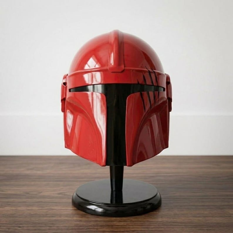K&ouml;nnte beinhalten: Ein gl&auml;nzender roter Helm mit einem T-f&ouml;rmigen schwarzen Visier und einem Mittelsteg, der von vorne nach hinten verl&auml;uft. Der Helm weist auf der rechten Seite schwarze kratzerartige Markierungen auf und steht auf einem runden schwarzen Sockel.
