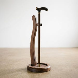 Könnte beinhalten: Ein dunkelbrauner Holzspazierstock mit gebogenem Griff und Messingakzenten, präsentiert in einem passenden Holzständer. Der Ständer hat eine kreisförmige Basis und eine gebogene Halterung. Der Stock ist etwa 91 cm hoch.