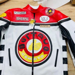 May include: A white, red, and black leather motorcycle jacket with a zip-up front. The jacket features patches with text and logos, including "PURSANG", "SHERPA", and "OTTADUO CEMOTO". The sleeves have black and white accents.