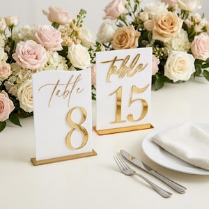May include: Two white rectangular acrylic table signs with gold mirrored bases. Each sign features the word Table in gold script lettering above a large gold mirrored number, specifically 8 and 15, standing upright on a smooth surface.