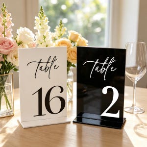 May include: Two rectangular acrylic table signs with stands. One sign is white with black text reading Table 16, and the other is black with white text reading Table 2. Both signs feature a script font for the word Table and large serif numerals.