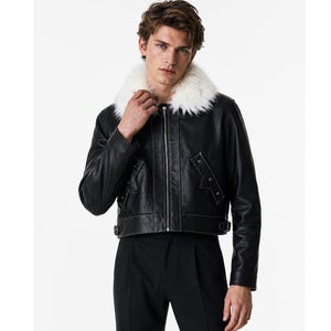 Pode incluir: Um casaco de couro preto com gola de pelo sintético branco, fecho de correr prateado à frente e dois bolsos laterais com detalhes de tachas prateadas. O casaco tem um corte curto com fivelas laterais ajustáveis na bainha e mangas.