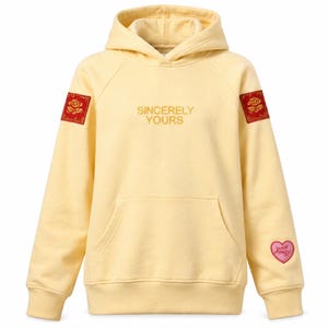 May include: A pale yellow hoodie with a front pocket and a hood. The text "Sincerely Yours" is embroidered on the chest. Red square patches with a floral design are on the shoulders, and a pink heart patch is on the sleeve.