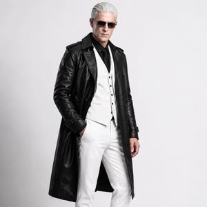 Pode incluir: Um sobretudo longo de couro preto usado sobre um colete branco, uma camisa preta com gravata preta e calças brancas. O traje inclui óculos de sol escuros e apresenta um design de peito duplo com dragonas nos ombros.