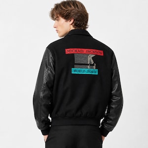 May include: Black bomber jacket with leather sleeves. The back of the jacket features a graphic with the text "MICHAEL JACKSON WORLD TOUR" in red and blue lettering. The jacket has a black collar and a black bottom hem.