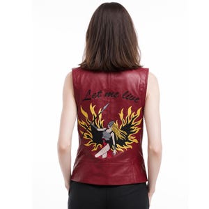 Op de afbeelding: Een mouwloos bordeauxrood vest met een geborduurde afbeelding op de achterkant. Het ontwerp toont een figuur met lang blond haar die een speer vasthoudt, omringd door gele en zwarte vlammen, met de tekst Let me live erboven geborduurd.