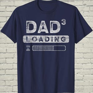 May include: Navy blue t-shirt with a distressed white graphic that reads "DAD 3 LOADING". The design includes a loading bar and an hourglass symbol, suggesting anticipation. A casual tee for a new dad.