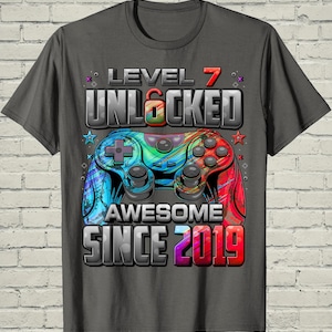 May include: Gray t-shirt with a colorful video game controller graphic. The design includes the text "LEVEL 7 UNLOCKED AWESOME SINCE 2019" in a bold, 3D style. The controller is a mix of blue, green, and red.