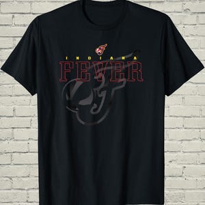 May include: A black short-sleeve t-shirt featuring the text INDIANA FEVER in red outlined lettering. Above the text is a small logo with a stylized letter F in red, yellow, and blue. A large, dark, semi-transparent graphic of the letter F is centered.