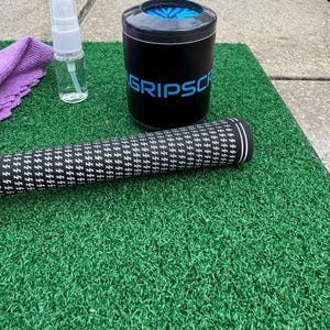 May include: A golf club grip cleaning kit on a green mat. The kit includes a black and white patterned grip, a black container with "GRIPSCR" in blue, a spray bottle, and a purple microfiber cloth.
