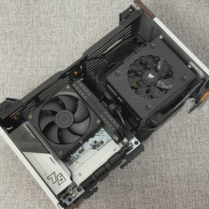 May include: A partially assembled computer case with various components visible. The case contains a black fan, a power supply unit, and a motherboard with other components. The components are black and silver, and the case is open, revealing the internal structure.