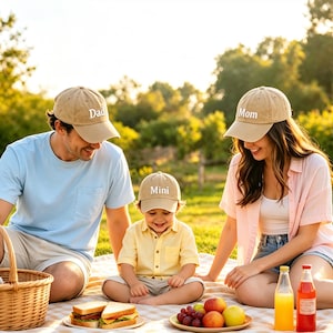 May include: A family picnic scene featuring tan baseball caps with the words "Dad", "Mom", and "Mini" in white. The picnic includes a basket, sandwiches, fruit, and drinks on a checkered blanket. The setting is a sunny outdoor area with trees in the background.