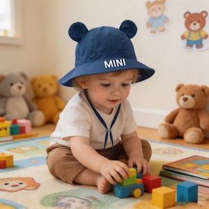 May include: A navy blue baby sun hat with bear ears and the word "MINI" in white. The child is wearing a white top and brown trousers, playing with colourful wooden blocks on a patterned rug. Teddy bears are in the background.