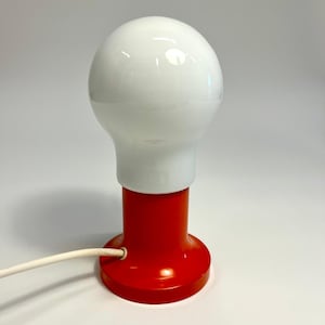 May include: A table lamp featuring a large, spherical white glass shade shaped like a light bulb, set upon a cylindrical, glossy red base. A white electrical cord extends from the side of the base, providing power to the lighting fixture.