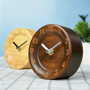 May include: Two small, round wooden clocks. One is light-colored, the other is dark brown. Both have visible numbers and black hands. The clocks are on a checkered surface, with a blue background and a hint of greenery.