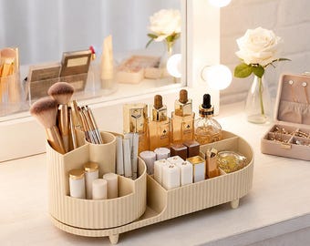 Peut inclure: Un organisateur de cosmétiques beige avec plusieurs compartiments, contenant des pinceaux de maquillage, des rouges à lèvres et des flacons. L'organisateur est posé sur une surface blanche, avec un miroir et des fleurs en arrière-plan. L'organisateur a un design côtelé.