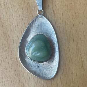 May include: A silver-toned pendant featuring an asymmetrical, brushed metal frame with a radial texture. Centered within the frame is a smooth, polished, rounded light green stone. The pendant includes a triangular bail at the top for hanging.
