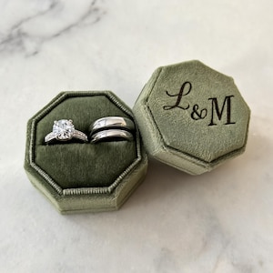 May include: An octagonal olive green velvet ring box with the letters L&M engraved on the lid. Inside the box are three silver-toned rings: one engagement ring with a central round-cut stone and two plain, smooth wedding bands.