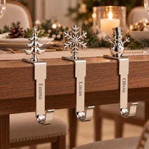 May include: Three silver-toned metal stocking holders with a polished finish. Each hook features a unique topper: a Christmas tree, a snowflake, and a snowman. The vertical stems are engraved with the names Emma, Lucas, and Olivia respectively.